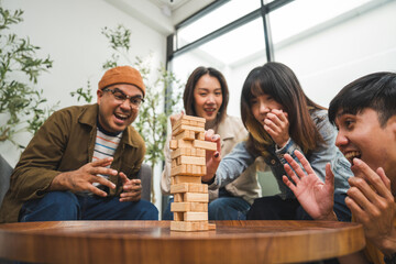 Young Asian friends are happily playing wooden block together at home, enjoying their leisure time. Group friends carefully remove a wooden block while others watch, having fun a classic table game.