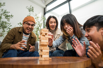 Young Asian friends are happily playing wooden block together at home, enjoying their leisure time. Group friends carefully remove a wooden block while others watch, having fun a classic table game.