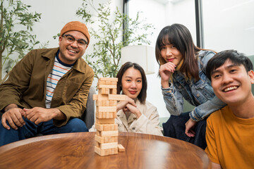 Young Asian friends are happily playing wooden block together at home, enjoying their leisure time. Group friends carefully remove a wooden block while others watch, having fun a classic table game.