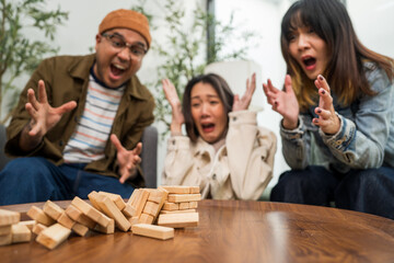 Young Asian friends are happily playing wooden block together at home, enjoying their leisure time. Group friends carefully remove a wooden block while others watch, having fun a classic table game.