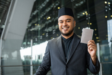 Muslim asian businessman traveler holding boarding pass ticket, passport and smartphone at the airport terminal with luggage. Cheerful tourist islamic at airport holiday trip.