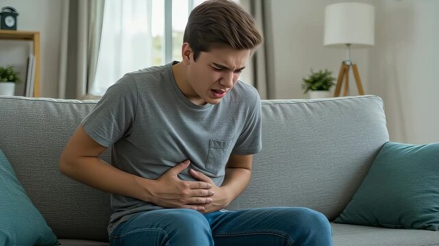 A young man sitting on a sofa at home, putting his hands on his stomach and complaining of stomach pain