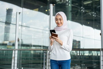 Fototapeta premium Muslim asian businesswoman traveler holding boarding pass ticket, passport and smartphone at the airport terminal with her luggage. Islamic cheerful tourist female having holiday trip weekend.