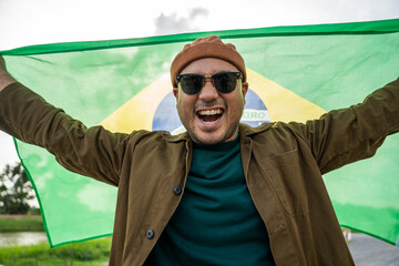 Portrait of cheerful young man looking forward while standing proudly with holding Brasil flag over...