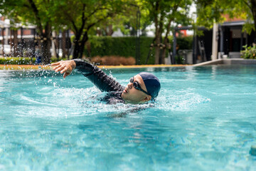 A person in a black rash guard and swim cap swims laps in a clear blue pool, demonstrating an...