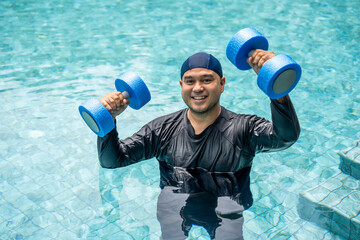 A smiling person in a swim cap and rash guard is submerged in a pool, holding blue aquatic...