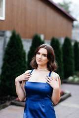 Girl in a long blue dress on the street in summer, green bushes behind her, she looks straight into...