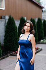 Girl in a long blue dress on the street in summer, green bushes behind her, she looks straight into...