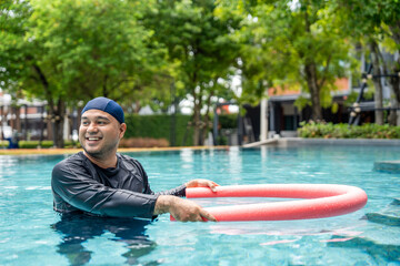 A smiling person in a swim cap and rash guard is in a pool, holding a pink swim noodle in an arc above their head. This image depicts aquatic exercise and joyful water recreation.