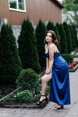 Girl in a long blue dress on the street in summer, green bushes behind her
