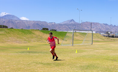 Male soccer player wearing red uniform sprinting on grass field dodging yellow cones by metal goal