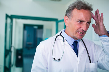 Senior male doctor raising hand to temple in hospital hall with teal doors stethoscope, copy space