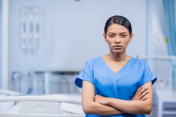 Female nurse standing confidently with arms crossed in hospital room with bed rails and IV pole