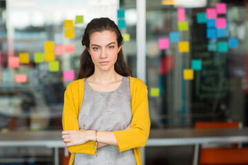 Woman wearing yellow cardigan crossing arms at office in front of partition with sticky notes