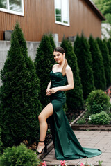 A girl in a green emerald dress walks down the street in summer, with large green bushes behind her.