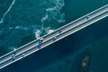 Aerial top down view of a cars driving along a coastal bridge with clear waters and ocean waves beneath in bright daylight