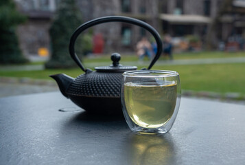 Glass cup with green tea and cast iron teapot on dark table, double wall glass
