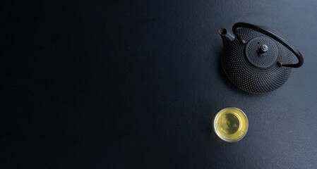 Glass cup with green tea and cast iron teapot on dark table, double wall glass