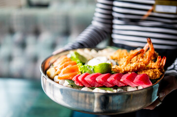 Close-up of a person holding a bowl of assorted seafood including raw fish, fried shrimp, lime, and greens, presented in a metal bowl with vibrant, appetizing detail.