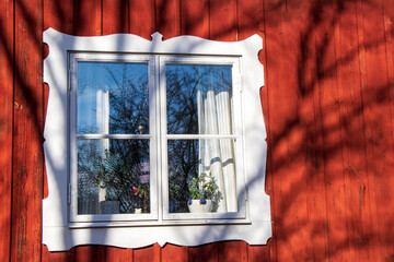 White-framed window on red wooden house in Stockholm with shadows and reflections.
Close-up of window with white frame on red wall reflecting trees and sky on sunny day in Stockholm.