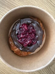 A close-up of a croissant in a paper bowl, topped with chocolate spread and a cherry filling, placed on a wooden surface.