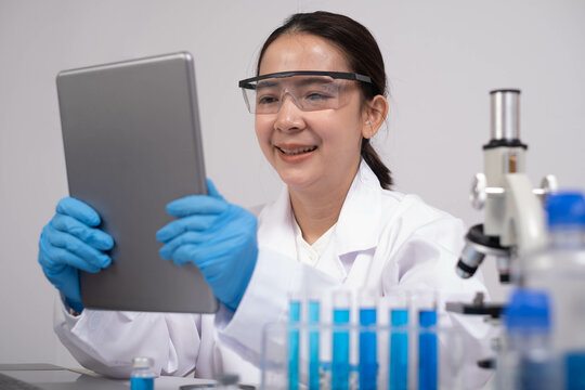A smiling woman in a lab coat and gloves uses a tablet while surrounded by scientific equipment and glassware.