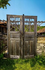 A wooden gate with two doors, one of which is locked. The gate is situated in a grassy area, surrounded by trees