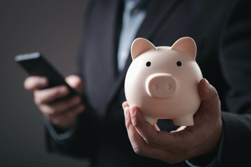 A business professional holds a piggy bank in one hand while using a smartphone, symbolizing...