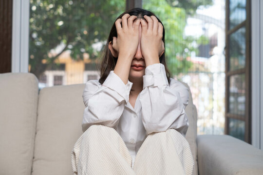 A woman sits on a couch, covering her face with her hands, expressing feelings of distress or overwhelm in a bright indoor setting.