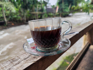 A cup of hot black coffee in a glass cup with matching saucer and spoon, placed on the edge of a...