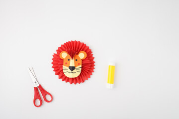 Lion face made from colored paper with red accordion mane placed between scissors and glue stick on white background. Flat lay composition with copy space. DIY paper animal project.