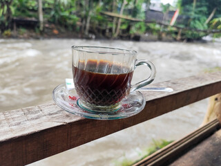A cup of hot black coffee in a glass cup with matching saucer and spoon, placed on the edge of a...