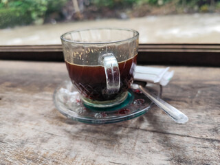 A cup of hot black coffee in a glass cup with matching saucer and spoon, placed on the edge of a weathered wooden fence with a backdrop of a flowing river and blurry green trees. Outdoor cafe menu.