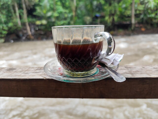 A cup of hot black coffee in a glass cup with matching saucer and spoon, placed on the edge of a weathered wooden fence with a backdrop of a flowing river and blurry green trees. Outdoor cafe menu.