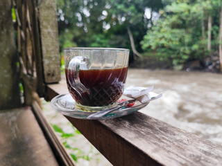 A cup of hot black coffee in a glass cup with matching saucer and spoon, placed on the edge of a weathered wooden fence with a backdrop of a flowing river and blurry green trees. Outdoor cafe menu.