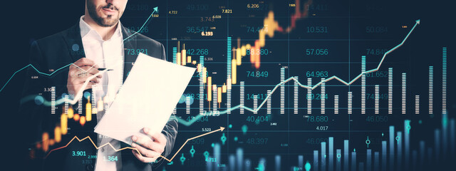 Businessman analyzing financial report with stock market holograms and data charts overlayed on...