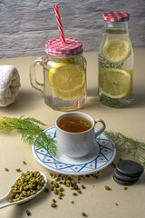 Fennel tea served in a white cup and saucer, accompanied by fennel seeds, lemon water in glass containers, a rolled towel, and black macarons on a light surface. Calm and healthy mood