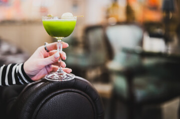 Close-up of a hand holding a green cocktail with a round white garnish, blurred cozy interior...