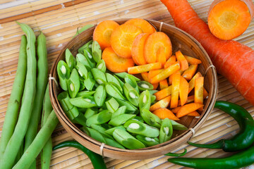Sliced green beans and fresh carrots are served in a bamboo basket on a bamboo mat