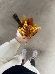 A person holding a cone of French fries topped with sauce, fried onions, and sausage, photographed from a top-down angle on a concrete background.