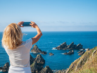 A woman photographs jagged sea stacks off the rugged coast of Galicia, Spain, under a bright summer...