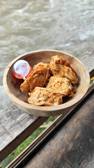 Tahu walik. Crispy browned tofu walik served in a natural wooden bowl with spicy chili sauce, with a blurry river flowing in the background. Menu at an outdoor cafe.