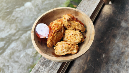 Tahu walik. Crispy browned tofu walik served in a natural wooden bowl with spicy chili sauce, with a blurry river flowing in the background. Menu at an outdoor cafe.