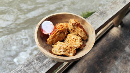 Tahu walik. Crispy browned tofu walik served in a natural wooden bowl with spicy chili sauce, with a blurry river flowing in the background. Menu at an outdoor cafe.