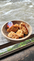 Tahu walik. Crispy browned tofu walik served in a natural wooden bowl with spicy chili sauce, with a blurry river flowing in the background. Menu at an outdoor cafe.