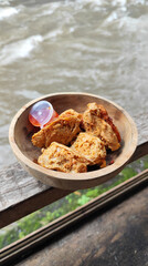 Tahu walik. Crispy browned tofu walik served in a natural wooden bowl with spicy chili sauce, with a blurry river flowing in the background. Menu at an outdoor cafe.