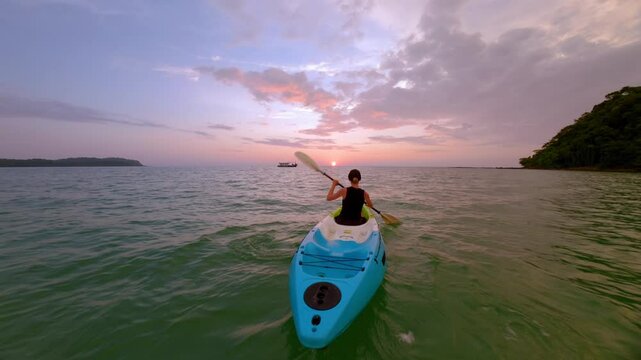 Woman kayaking on calm turquoise sea during golden sunset in Thailand. Peaceful tropical scenery with colorful sky, ideal for travel, freedom, and adventure themes.