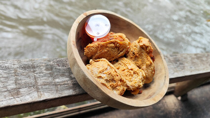 Tahu walik. Crispy browned tofu walik served in a natural wooden bowl with spicy chili sauce, with a blurry river flowing in the background. Menu at an outdoor cafe.