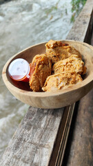 Tahu walik. Crispy browned tofu walik served in a natural wooden bowl with spicy chili sauce, with a blurry river flowing in the background. Menu at an outdoor cafe.