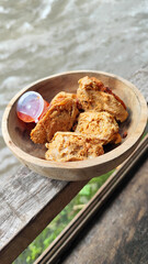 Tahu walik. Crispy browned tofu walik served in a natural wooden bowl with spicy chili sauce, with a blurry river flowing in the background. Menu at an outdoor cafe.